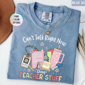 May include: Blue jean colored t-shirt with the text "Can't Talk Right Now Doing Teacher Stuff". The graphic includes a notepad, pencil, apple, and a pink tumbler. A gold necklace is draped across the shirt.
