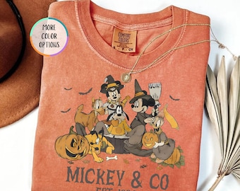 Op de afbeelding: Een yamkleurig T-shirt met lange mouwen met een Halloween-thema van Mickey Mouse en vrienden. De afbeelding bevat pompoenen, vleermuizen en de tekst "Mickey & Co. Est. 1928". Een gouden ketting ligt op het shirt.