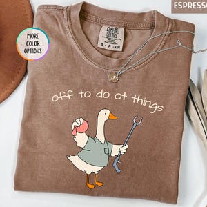 May include: An espresso-colored short-sleeve t-shirt featuring a graphic of a white goose wearing a green shirt, holding a pink object and a reacher grabber tool. The text "off to do ot things" is printed above the goose in a simple white font.
