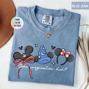 May include: Blue jean colored t-shirt with a graphic featuring Mickey Mouse ears, a wizard hat, and Minnie Mouse ears. The shirt has the text "Some imagination, huh?" and a gold necklace. The shirt is labeled "Comfort Colors".