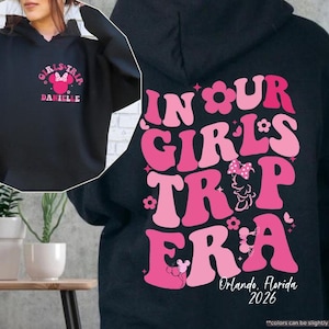 Peut inclure: Sweat &agrave; capuche noir avec un motif rose au dos indiquant "IN OUR GIRLS TRIP ERA" avec un design Minnie Mouse et le texte "Orlando, Florida 2026". L'avant du sweat &agrave; capuche porte un motif plus petit avec "GIRLS TRIP" et "DANIELLE".