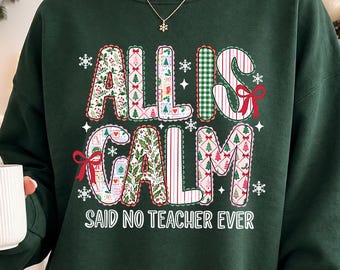 以下が含まれることがあります： クリスマスモチーフの柄が入った大きなブロック体で「ALL IS CALM」と書かれ、その下に白いサンセリフ体の大文字で「SAID NO TEACHER EVER」とプリントされた、ダークグリーンの長袖スウェットシャツ。