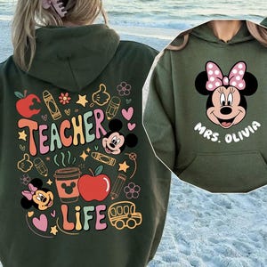 May include: An olive green hoodie with a graphic of Minnie Mouse and the text "MRS. OLIVIA" on the front. The back features a colorful design with the words "TEACHER LIFE", apples, pencils, and Mickey Mouse.