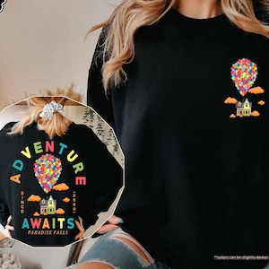 Puede incluir: Sudadera negra con un gr&aacute;fico colorido de una casa levantada por globos, con el texto "Adventure Awaits Paradise Falls" y el a&ntilde;o "2009". El dise&ntilde;o se repite en la espalda. La sudadera es negra.