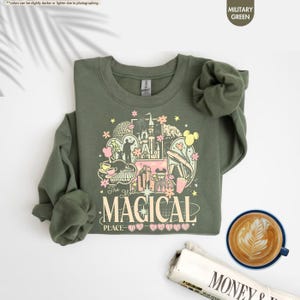 May include: A military green sweatshirt with a colorful graphic design featuring a castle, Mickey Mouse ears, and the text "The Most Magical Place". The sweatshirt is folded, with a cup of coffee and a newspaper in the background.