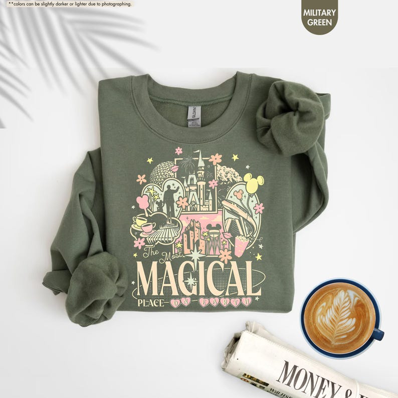 May include: A military green sweatshirt with a colorful graphic design featuring a castle, Mickey Mouse ears, and the text "The Most Magical Place". The sweatshirt is folded, with a cup of coffee and a newspaper in the background.