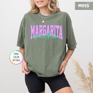May include: A moss green t-shirt with the words "MARGARITA weather" in a pink and teal font. The shirt is a relaxed fit and has short sleeves. The image also shows a person wearing the shirt.