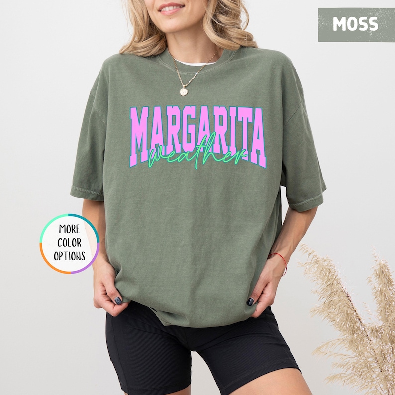 May include: A moss green t-shirt with the words "MARGARITA weather" in a pink and teal font. The shirt is a relaxed fit and has short sleeves. The image also shows a person wearing the shirt.