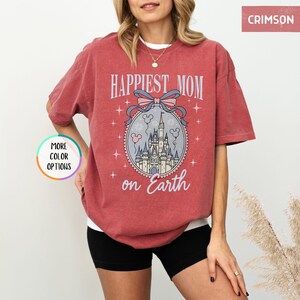 May include: Crimson-colored t-shirt with the text "Happiest Mom on Earth" and a graphic of a castle, bow, and Mickey Mouse balloons. The shirt has short sleeves and a relaxed fit.