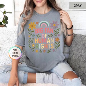 May include: Gray t-shirt with the text "BIG FAN OF HUMAN RIGHTS" in colorful letters, surrounded by floral and rainbow graphics. The shirt is a casual style, suitable for everyday wear.