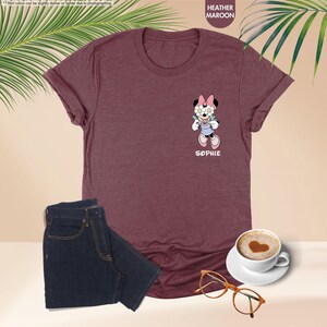 May include: Heather maroon t-shirt with a Minnie Mouse graphic wearing flower glasses and overalls. The name "SOPHIE" is printed below the graphic. A pair of dark blue jeans, a cup of coffee, and glasses are also in the image.