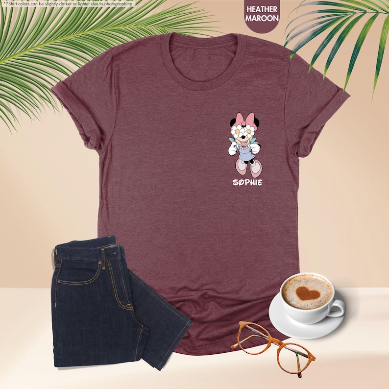 May include: Heather maroon t-shirt with a Minnie Mouse graphic wearing flower glasses and overalls. The name "SOPHIE" is printed below the graphic. A pair of dark blue jeans, a cup of coffee, and glasses are also in the image.