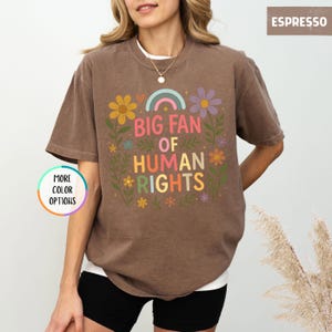 May include: Espresso-colored t-shirt with the text "BIG FAN OF HUMAN RIGHTS" in colorful letters and floral accents. The shirt has a crew neck and short sleeves. The words "MORE COLOR OPTIONS" are also visible.
