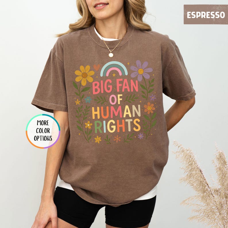 May include: Espresso-colored t-shirt with the text "BIG FAN OF HUMAN RIGHTS" in colorful letters and floral accents. The shirt has a crew neck and short sleeves. The words "MORE COLOR OPTIONS" are also visible.
