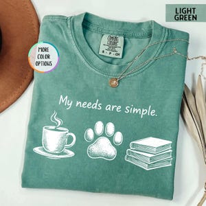 May include: Light green t-shirt with the phrase "My needs are simple." and illustrations of a coffee cup, paw print, and books. The shirt has a crew neck and short sleeves. A gold necklace is draped across the shirt.