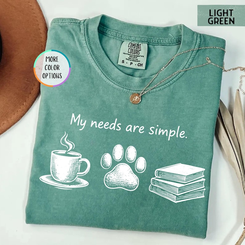 May include: Light green t-shirt with the phrase "My needs are simple." and illustrations of a coffee cup, paw print, and books. The shirt has a crew neck and short sleeves. A gold necklace is draped across the shirt.