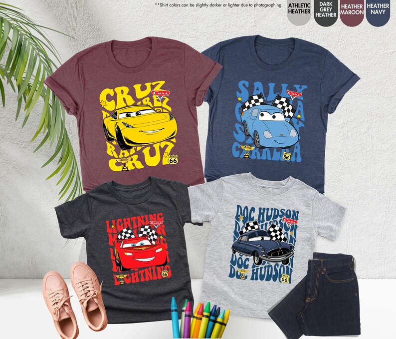 May include: Four children's t-shirts with Cars movie characters. The shirts are in shades of maroon, navy, dark grey, and athletic heather. Each shirt features a different car character with text and checkered flags, evoking a racing theme.