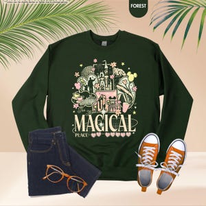 May include: Forest green sweatshirt with a colorful graphic that reads "The Most Magical Place." The graphic includes a castle, Mickey Mouse, and other theme park elements. Paired with blue jeans, orange sneakers, and glasses.