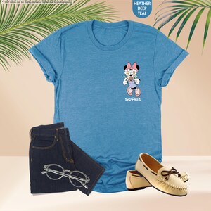 May include: A heather deep teal t-shirt featuring a Minnie Mouse graphic holding flowers, with the name "Sophie" below. The shirt is paired with dark blue jeans, eyeglasses, and cream-colored moccasins.