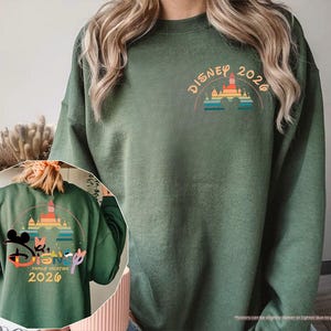 May include: Military green sweatshirt with a colorful Disney castle graphic and the text "DISNEY 2026" on the front. The back of the sweatshirt features a larger graphic with Mickey and Minnie Mouse, the text "FAMILY VACATION 2026", and the Disney logo.