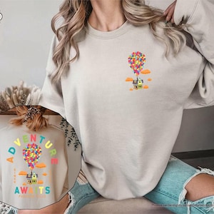 Puede incluir: Sudadera de color arena con un gr&aacute;fico colorido de una casa levantada por globos, con el texto "Adventure Awaits". El dise&ntilde;o incluye nubes naranjas y un grupo de globos multicolores. La sudadera es de color neutro.