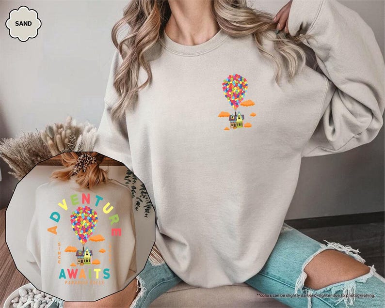 Puede incluir: Sudadera de color arena con un gr&aacute;fico colorido de una casa levantada por globos, con el texto "Adventure Awaits". El dise&ntilde;o incluye nubes naranjas y un grupo de globos multicolores. La sudadera es de color neutro.