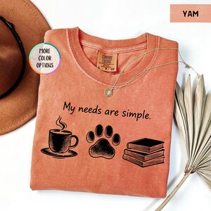 May include: A yam-colored t-shirt with the phrase "My needs are simple" printed in black. The design includes a coffee cup, paw print, and books. A gold necklace rests on the shirt.