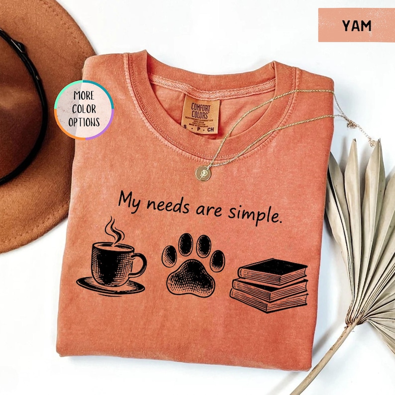 May include: A yam-colored t-shirt with the phrase "My needs are simple" printed in black. The design includes a coffee cup, paw print, and books. A gold necklace rests on the shirt.
