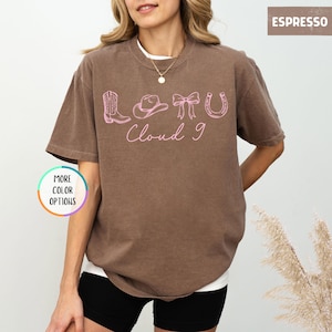 May include: Espresso-colored t-shirt with a white undershirt. The shirt features a pink graphic of a cowboy boot, hat, bow, horseshoe, and the words "Cloud 9". A button on the left says "More Color Options".