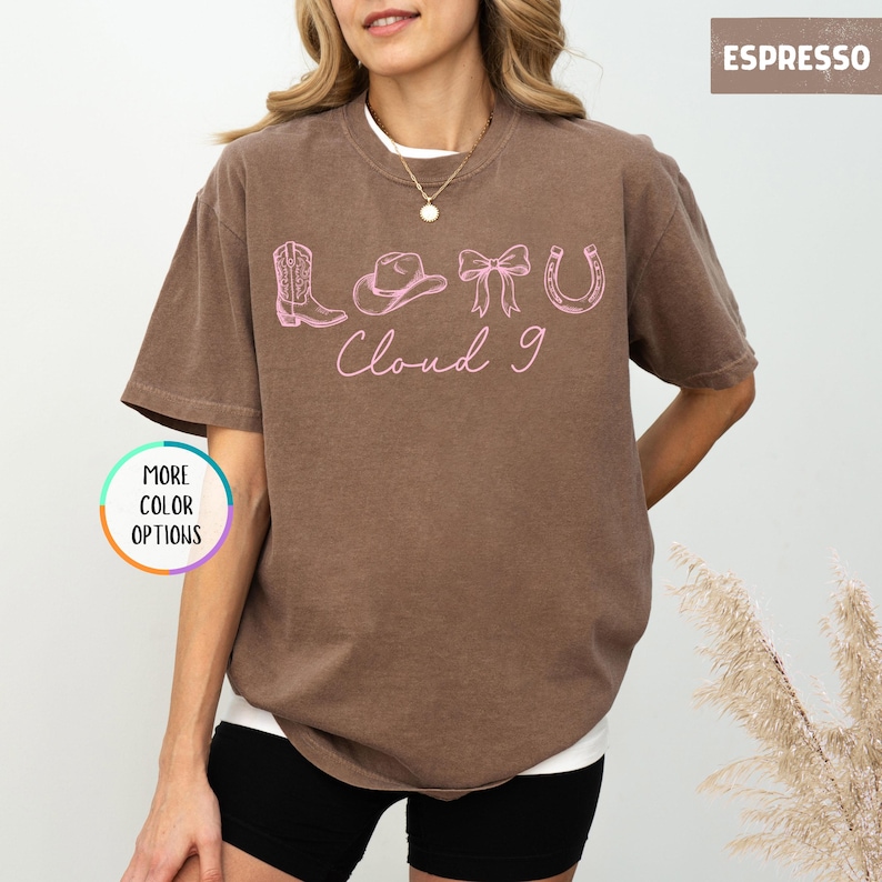 May include: Espresso-colored t-shirt with a white undershirt. The shirt features a pink graphic of a cowboy boot, hat, bow, horseshoe, and the words "Cloud 9". A button on the left says "More Color Options".