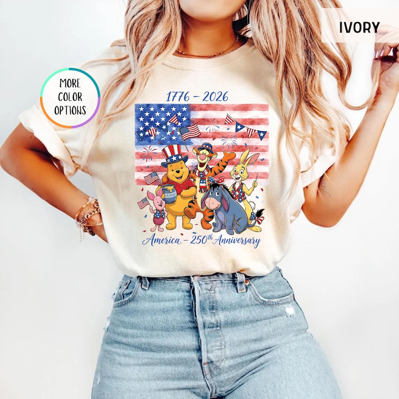 May include: Ivory-colored t-shirt featuring a graphic of Winnie the Pooh and friends in front of an American flag. The design includes the text "1776-2026" and "America - 250th Anniversary". The shirt has short sleeves and a round neckline.