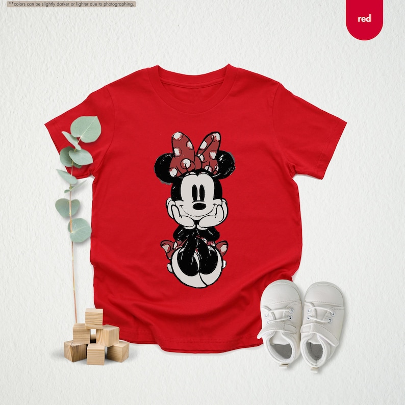 May include: A red t-shirt featuring a Minnie Mouse graphic. The cartoon character is depicted in black and white with a red bow. White baby shoes are placed next to the shirt.