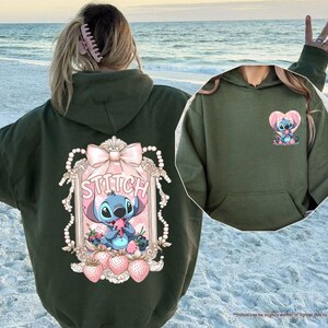 Puede incluir: Sudadera con capucha verde oscuro con un gr&aacute;fico de Stitch en la espalda y un dise&ntilde;o de coraz&oacute;n de Stitch m&aacute;s peque&ntilde;o en la parte delantera. El gr&aacute;fico de la espalda incluye la palabra "Stitch" en un marco decorativo con un lazo, perlas y fresas.