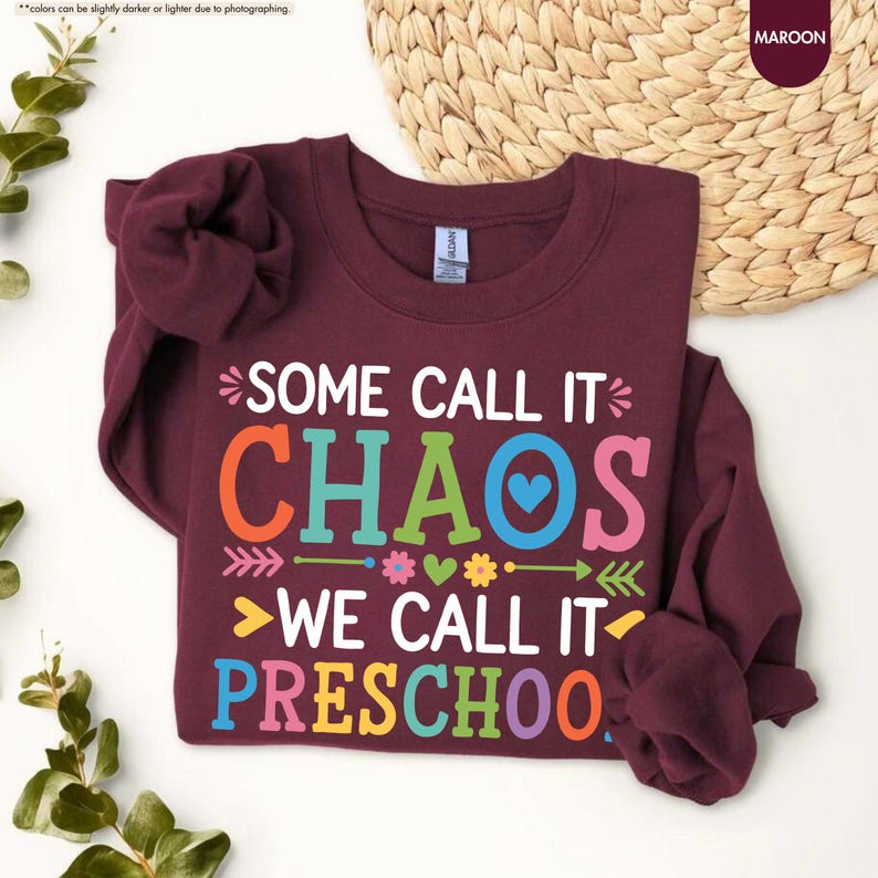 May include: Maroon sweatshirt with the text "SOME CALL IT CHAOS WE CALL IT PRESCHOOL" in colorful letters. The design includes arrows, hearts, and flowers. The sweatshirt is displayed on a white surface with a woven basket in the background.