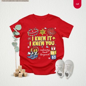 Peut inclure: Un t-shirt rouge &agrave; manches courtes avec une impression graphique portant le texte I KNEW IT I KNEW YOU infinity and beyond. Le motif comprend un insigne de sh&eacute;rif, un chapeau de cow-boy, un bracelet de perles, des bottes et trois cubes.
