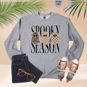 May include: A sport grey crewneck sweatshirt featuring the black text SPOOKY SEASON and three ghost illustrations with camouflage, leopard print, and checkerboard patterns. The garment is displayed with dark denim jeans, orange sneakers, and glasses.