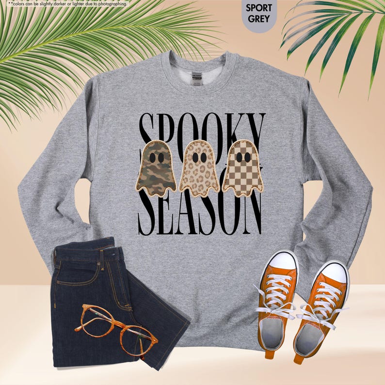 May include: A sport grey crewneck sweatshirt featuring the black text SPOOKY SEASON and three ghost illustrations with camouflage, leopard print, and checkerboard patterns. The garment is displayed with dark denim jeans, orange sneakers, and glasses.