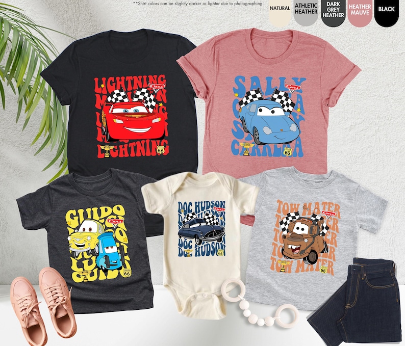 May include: A selection of children's t-shirts and a onesie with cartoon car designs. The shirts are in various colours, such as black, pink, and grey. The designs feature the text "Lightning McQueen", "Sally Carrera", "Guido", "Doc Hudson", and "Tow Mater".