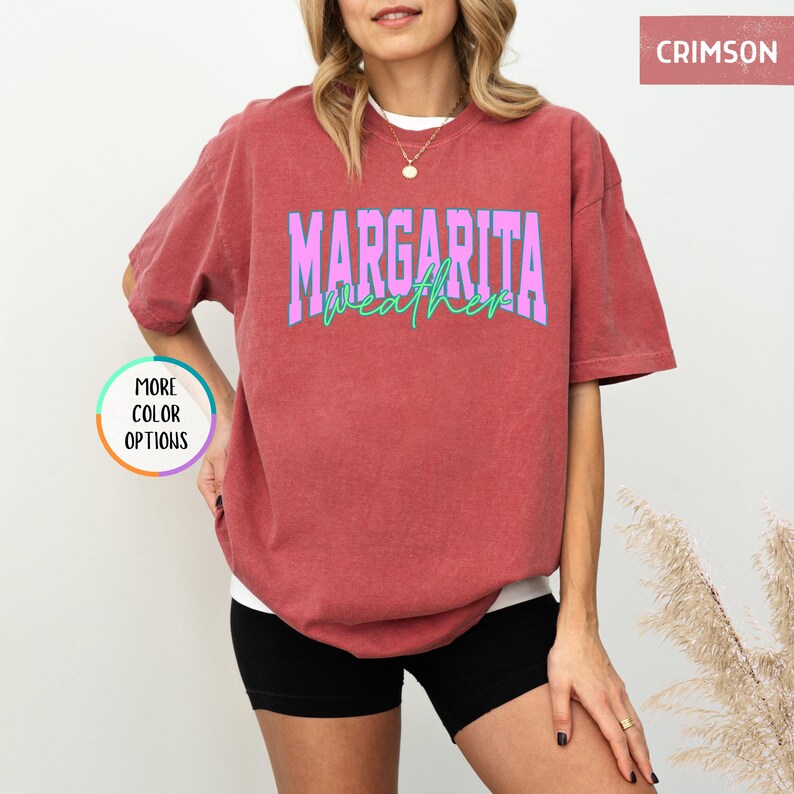 May include: Crimson-colored t-shirt with the text "MARGARITA weather" in a two-tone design. The word "MARGARITA" is in purple with a light blue outline, and "weather" is in light green. The shirt is a relaxed fit.