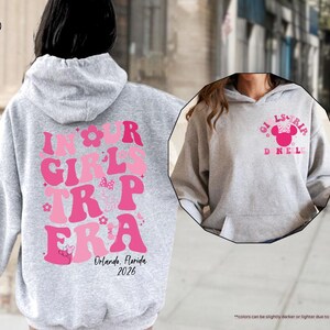 Peut inclure: Un sweat &agrave; capuche gris clair avec du texte rose qui dit "IN OUR GIRLS TRIP ERA" avec "Orlando, Florida 2026" en dessous. Le devant du sweat &agrave; capuche a un motif Minnie Mouse avec "GIRLS TRIP DNEL" en rose. Le sweat &agrave; capuche est de couleur gris sport.