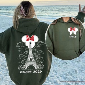 May include: A military green hooded sweatshirt featuring a white graphic of the Eiffel Tower, clouds, and hearts on the back with the text Disney 2026. The front chest area displays a white Minnie Mouse silhouette with a red polka dot bow and the name Emily.