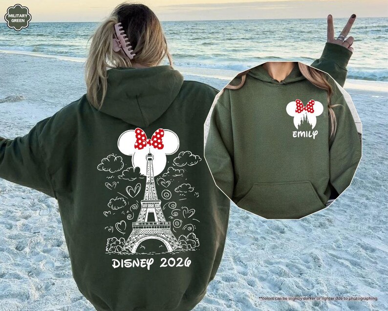 May include: A military green hooded sweatshirt featuring a white graphic of the Eiffel Tower, clouds, and hearts on the back with the text Disney 2026. The front chest area displays a white Minnie Mouse silhouette with a red polka dot bow and the name Emily.