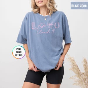May include: A blue jean colored t-shirt with a pink graphic that says "Love Cloud 9" with cowboy boot, hat, bow, and horseshoe icons. The shirt is a relaxed fit and has short sleeves.