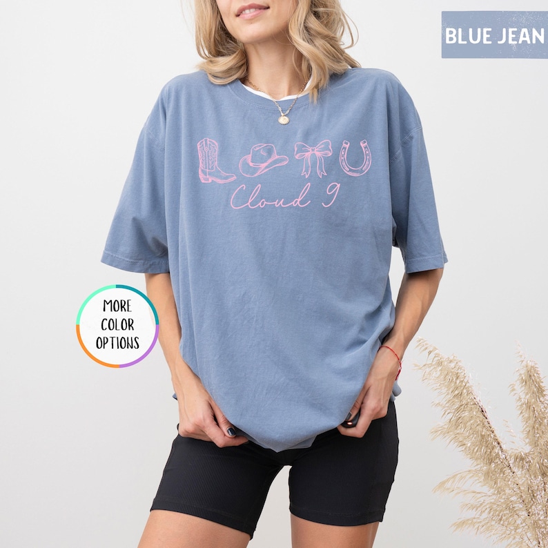 May include: A blue jean colored t-shirt with a pink graphic that says "Love Cloud 9" with cowboy boot, hat, bow, and horseshoe icons. The shirt is a relaxed fit and has short sleeves.