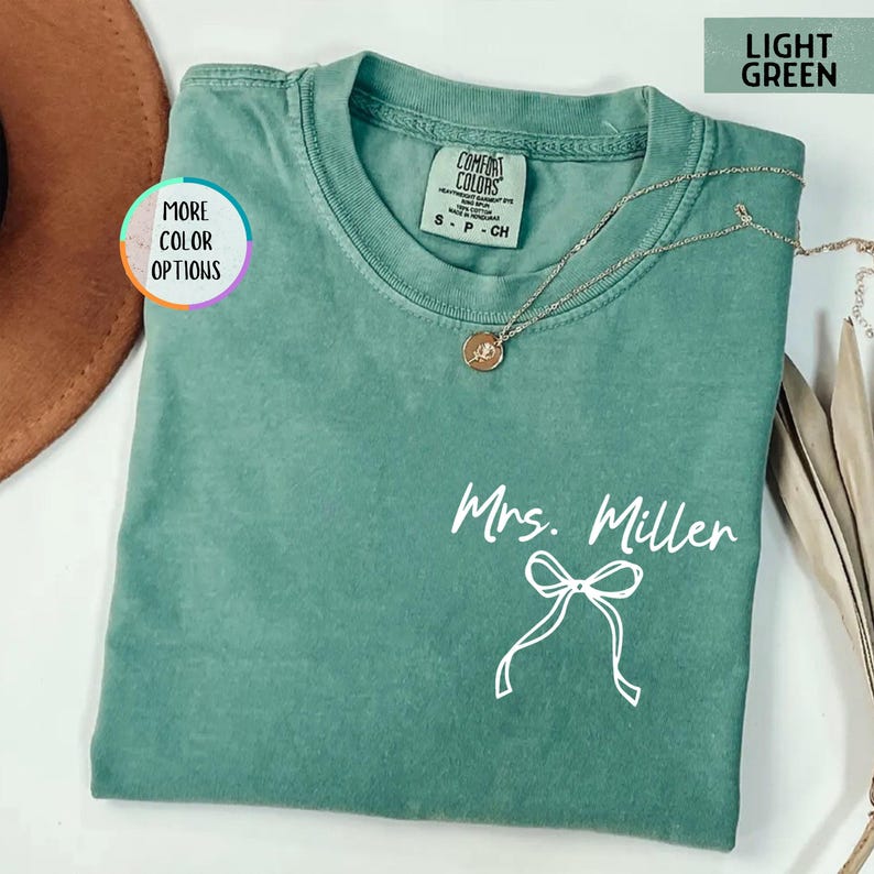 Puede incluir: Camiseta Comfort Colors verde claro con el texto "Mrs. Miller" y un lazo blanco. Un collar dorado con un colgante est&aacute; drapeado sobre la camiseta. La camiseta es talla peque&ntilde;a.