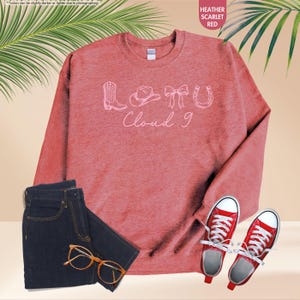 May include: A heather scarlet red sweatshirt with the text "Cloud 9" and cowboy-themed graphics. The image also includes a pair of dark wash jeans, red trainers, and gold-rimmed glasses. The sweatshirt is a casual, everyday wear item.