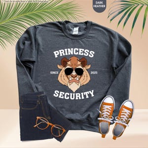 May include: Dark heather grey sweatshirt with a white graphic of a cartoon Beast from Beauty and the Beast wearing sunglasses. The graphic says "Princess Security" with the words "Since" and "2025" on either side of the graphic.