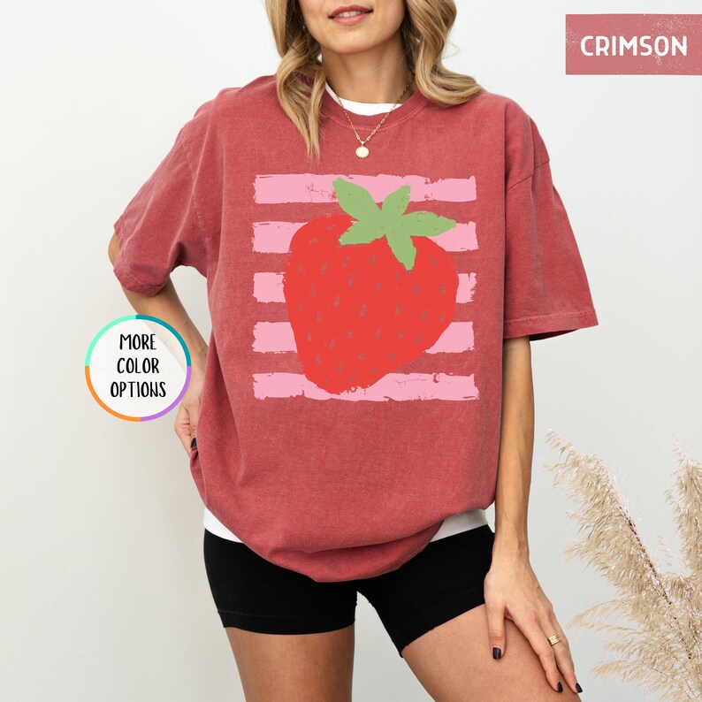 May include: A crimson short-sleeve t-shirt featuring a graphic of a red strawberry with a green stem centered over horizontal light pink brushstroke stripes. The text CRIMSON appears in the top right corner and MORE COLOR OPTIONS is on the left.