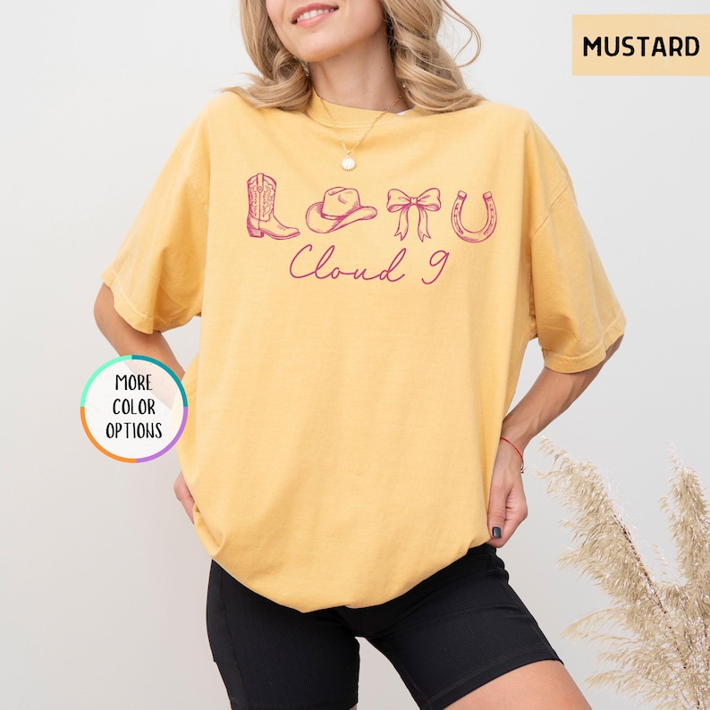 May include: Mustard-colored t-shirt with a pink graphic of a cowboy boot, hat, bow, and horseshoe, with the text "Cloud 9". The shirt has a relaxed fit and is worn by a person.