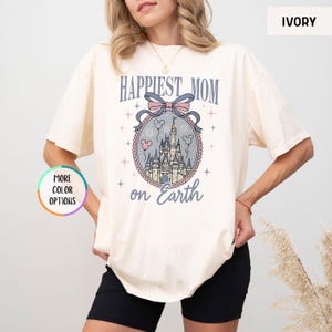 May include: Ivory-colored t-shirt with the text "Happiest Mom on Earth" above a castle graphic. The graphic includes a bow, stars, and Mickey Mouse balloons. The shirt is a relaxed fit.
